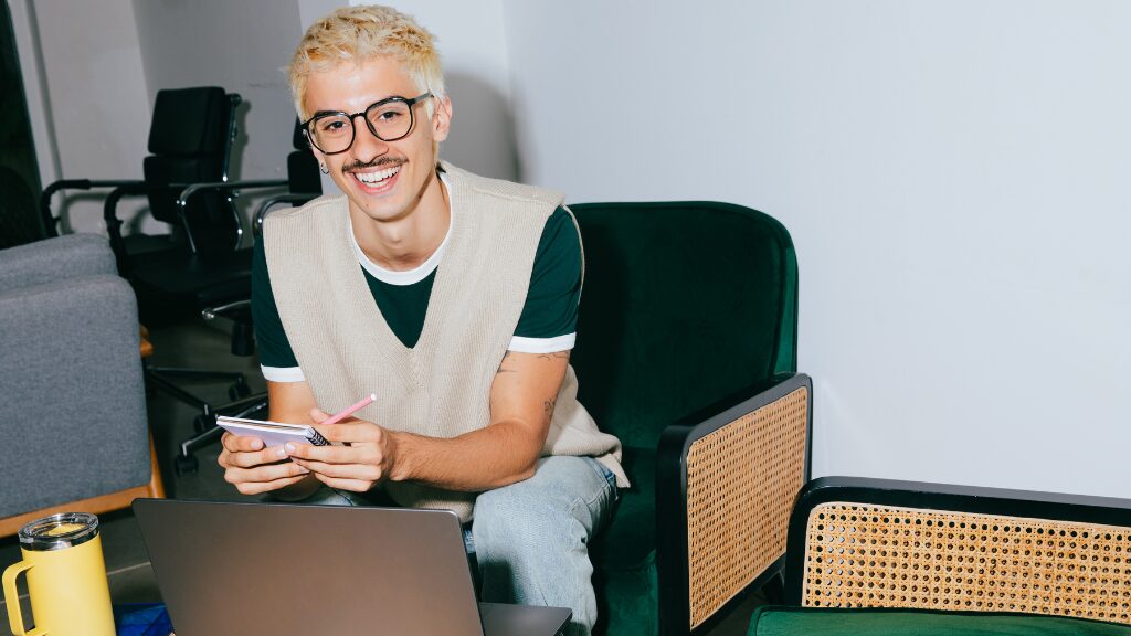 Étudiant souriant assis avec un ordinateur portable, prenant des notes dans un espace de travail moderne.